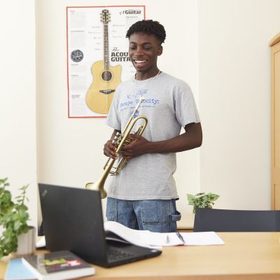 A boy is stood up, holding a trumpet and looking at a laptop